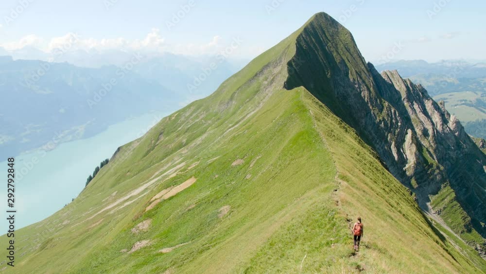 Female walking away in slow motion on dramatic ridge line of Swiss Alps hiking trail with lake in background and peak of mountain in distance Female walking away in slow motion on dramatic ridge line of Swiss Alps hiking trail with lake in background and peak of mountain in distance