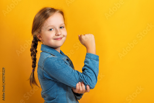 Closeup portrait of a little girl on a yellow background. Children's hand shows biceps. Girl power concept.