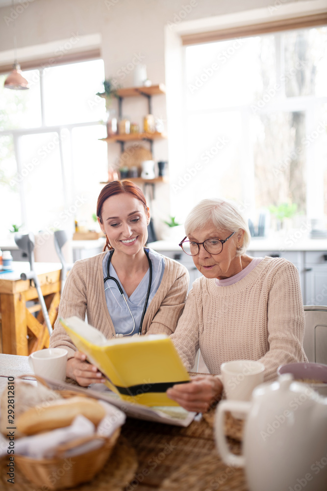 Nurse sitting near pensioner reading book in the morning Nurse sitting near pensioner reading book in the morning
