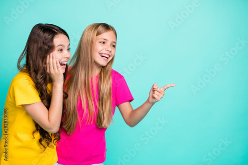 Photo of two nice cute cheerful girls with one of them pointing at emptiness and second being surprised while isolated with teal background with copyspace