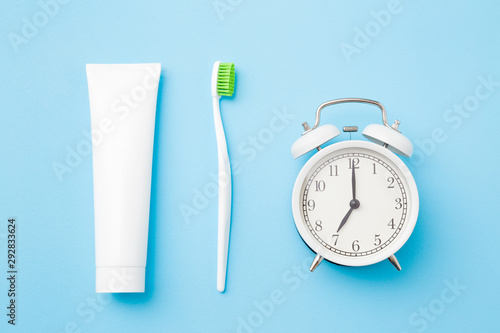 Alarm clock, toothbrush with green bristles and white tube of toothpaste on pastel blue background. Healthy teeth. Morning routine concept.  photo