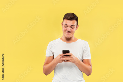 Caucasian young man's half-length portrait on yellow studio background. Beautiful male model in shirt. Concept of human emotions, facial expression, sales, ad. Using phone, looks crazy happy. © master1305