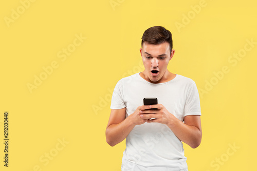 Caucasian young man's half-length portrait on yellow studio background. Beautiful male model in shirt. Concept of human emotions, facial expression, sales, ad. Using phone, looks shocked and upset.