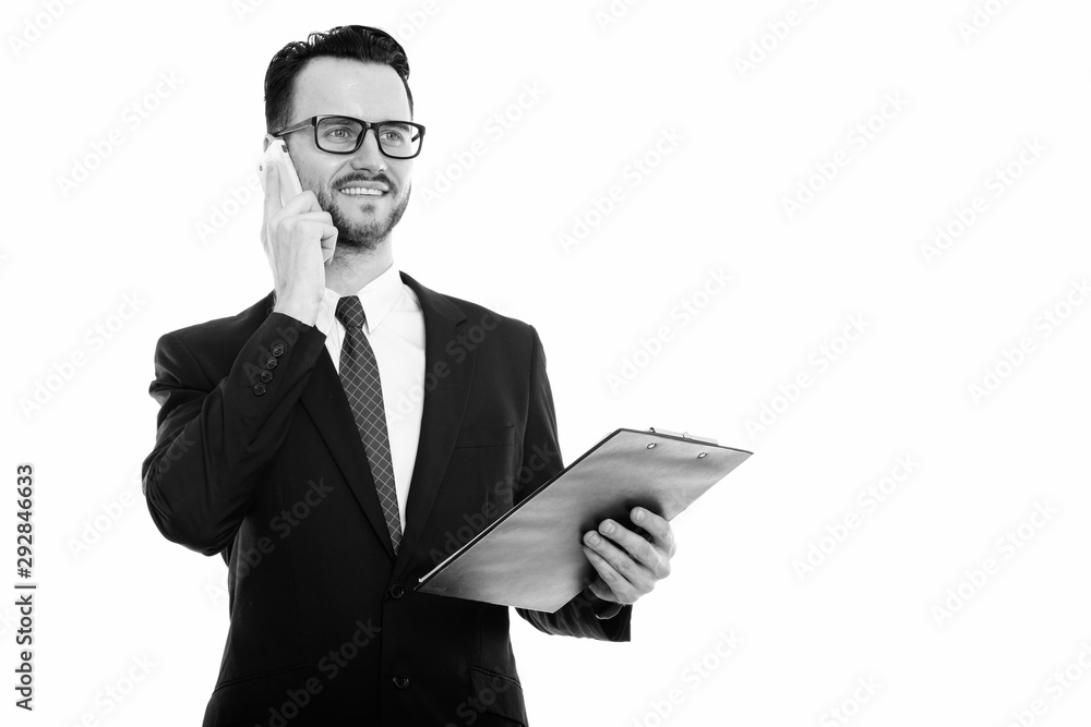 Studio shot of young happy businessman smiling while holding clipboard and talking on mobile phone Studio shot of young happy businessman smiling while holding clipboard and talking on mobile phone