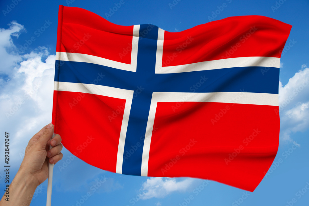 Fototapeta premium hand holds against the background of the sky with clouds the colored flag of Norway on the texture of the fabric, silk with waves, close-up