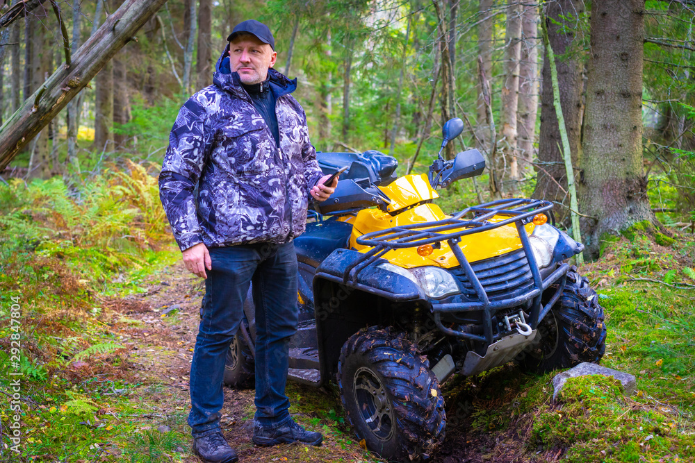 A man with a smartphone stands next to a yellow ATV. A man is going to take a photo in the woods. Outdoor activities. Walk through the woods on ATV. A man with a smartphone stands next to a yellow ATV. A man is going to take a photo in the woods. Outdoor activities. Walk through the woods on ATV.