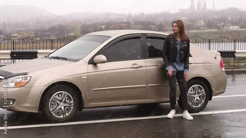 Portrait of a frustrated girl on an empty road in the rain near her broken car, she tries to call someone and catch passing cars. Portrait of a frustrated girl on an empty road in the rain near her broken car, she tries to call someone and catch passing cars.