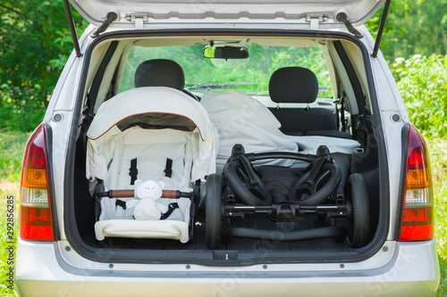 Parts of baby carriage in opened car trunk. Preparing to become parent for future child. Things transportation. Green outdoor nature and sunny summer day atmosphere.
