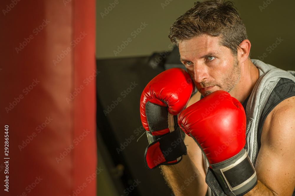 gym workout portrait of attractive and fierce looking boxer man 30s to 40s in boxing gloves training at fitness club punching  heavy bag in cool and badass fighter look