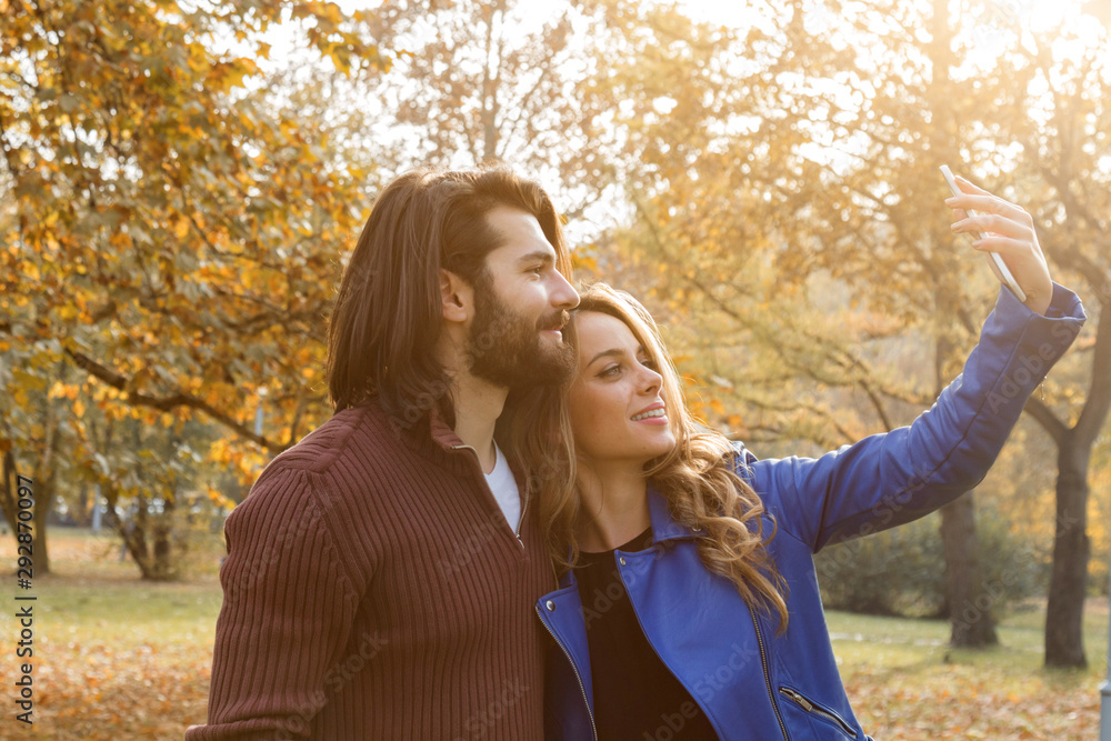 Fototapeta premium Young couple using cellphone in autumn colored park.