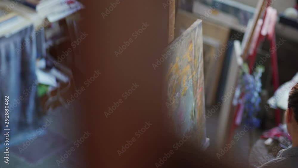 High angle tracking shot of inspired female artist using palette knife to paint abstract portrait on canvas standing at easel in workshop, side view High angle tracking shot of inspired female artist using palette knife to paint abstract portrait on canvas standing at easel in workshop, side view
