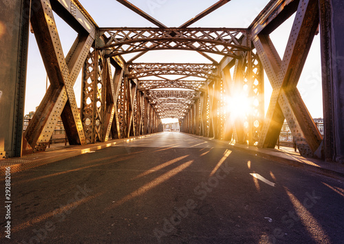 Fototapeta Naklejka Na Ścianę i Meble -  Asphalt road under the steel construction of a bridge in the city on a sunny day. Evening urban scene with the sunbeam in the tunnel. City life, transport and traffic concept.