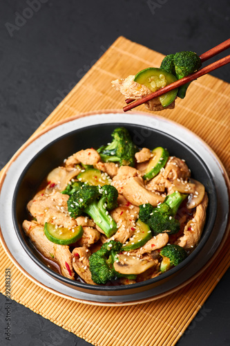 Hunan Chicken in black bowl at dark slate background. Hunan Chicken is chinese or indo-chinese cuisine takeaway dish with broccoli, zucchini, shiitake mushrooms and hunan sauce