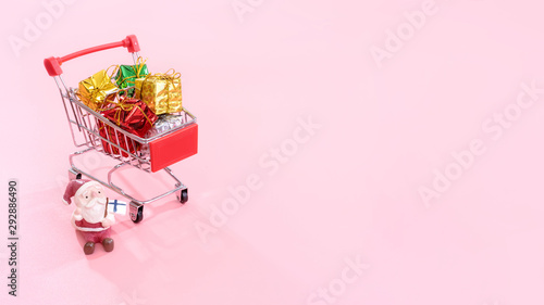 Christmas shopping concept, mini red shop cart trolley with Santa Claus toy and gift box isolated on pale pink background, blank copy space, close up