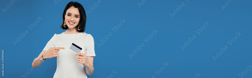 panoramic shot of smiling elegant woman in dress pointing with finger at credit card isolated on blue panoramic shot of smiling elegant woman in dress pointing with finger at credit card isolated on blue