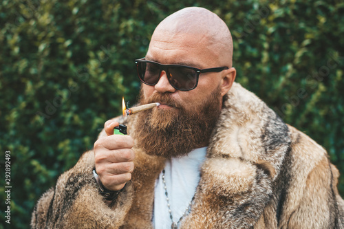 Portrait of posh chic virile bearded brutal man smoking marijuana joint, wearing brown fur gypsy style - hip hop pimp stylish guy lighting up weed (cannabis) blunt at the green background outdoors photo