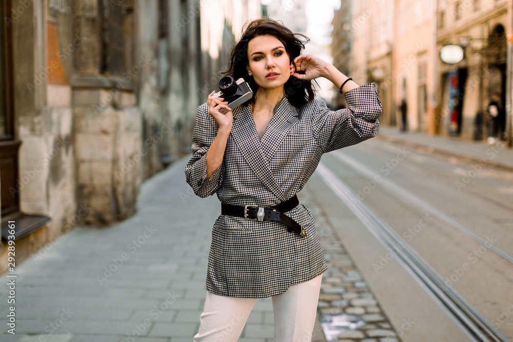 Attractive modern woman standing on the city street. Fashion portrait of pretty young woman in white pants and checkered jacket, holding old camera in hands