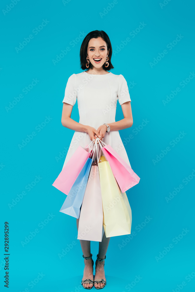 full length view of smiling elegant woman in dress with shopping bags isolated on blue full length view of smiling elegant woman in dress with shopping bags isolated on blue