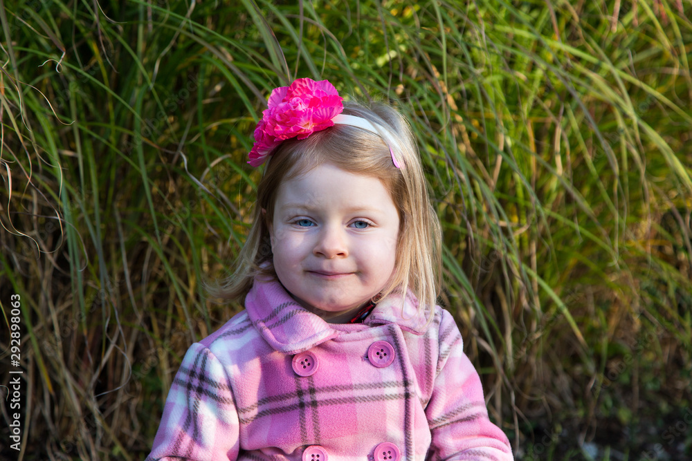 Medium horizontal portrait of pretty blue-eyed blond four-year old girl with happy expression in pink, brown and white checkered coat staring ahead while outdoors in the fall Medium horizontal portrait of pretty blue-eyed blond four-year old girl with happy expression in pink, brown and white checkered coat staring ahead while outdoors in the fall