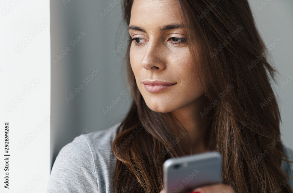 Close up of a young beautiful girl standing leaning on a gray wall over gray background, using mobile phone Close up of a young beautiful girl standing leaning on a gray wall over gray background, using mobile phone