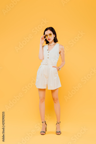 full length view of elegant woman in sunglasses posing with hand in pocket isolated on yellow