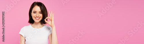 panoramic shot of smiling pretty girl showing okay sign isolated on pink photo