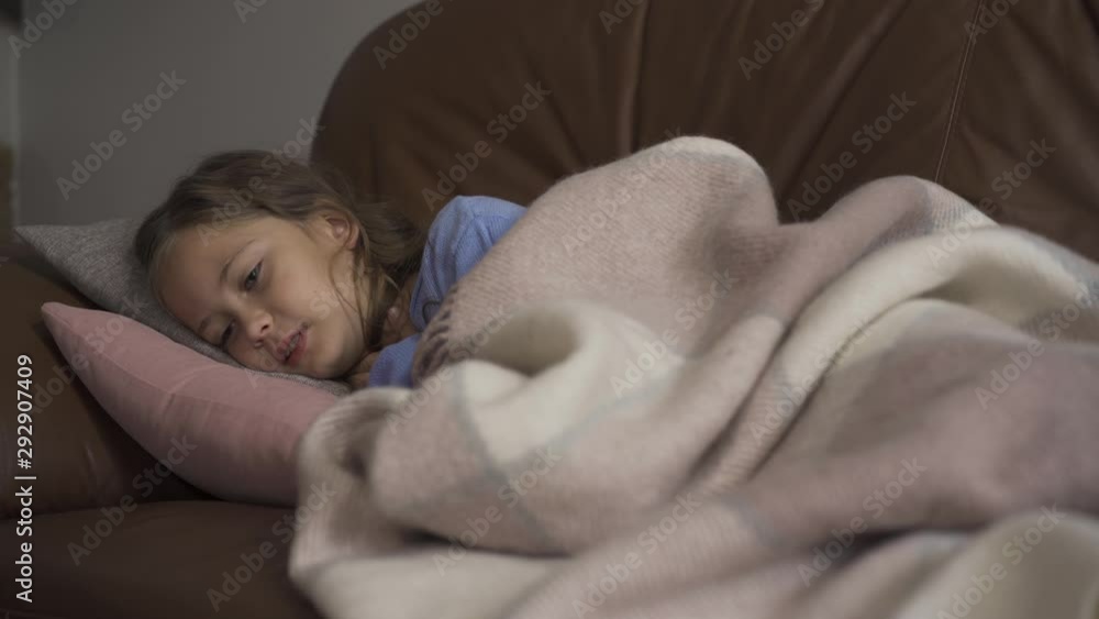 Sick young caucasian girl coughing while lying under blanket at home. The child has fever. Concept of health, illness, sickness, common cold, treatment Sick young caucasian girl coughing while lying under blanket at home. The child has fever. Concept of health, illness, sickness, common cold, treatment