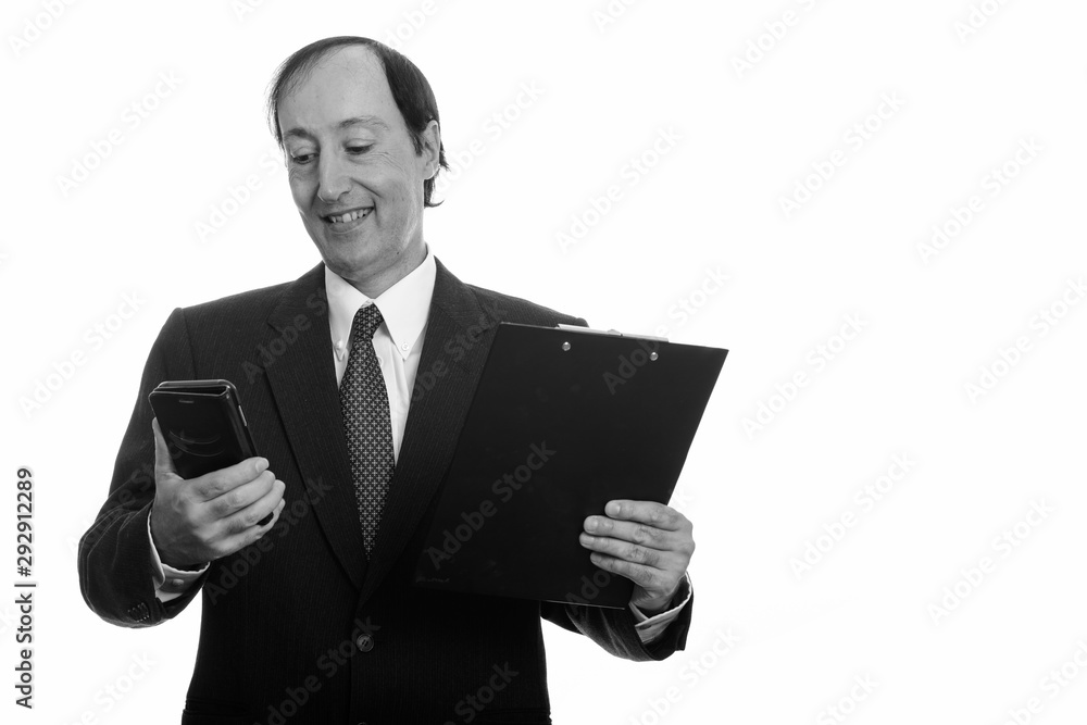 Studio shot of happy mature businessman smiling and using mobile phone while holding clipboard Studio shot of happy mature businessman smiling and using mobile phone while holding clipboard