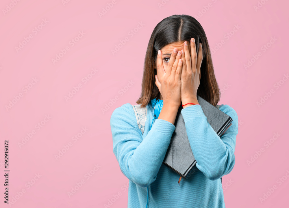 Young student girl with blue sweater and headphones covering eyes by hands and looking through the fingers on pink background Young student girl with blue sweater and headphones covering eyes by hands and looking through the fingers on pink background