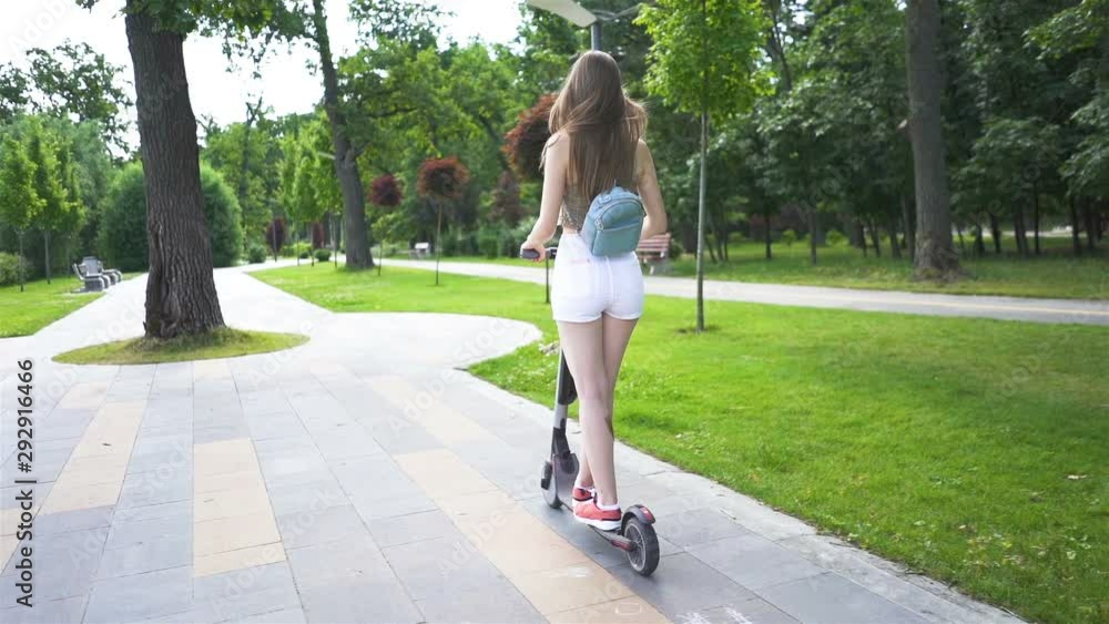 Slow motion. Young sexy girl riding electric scooter in city park area. Back view Slow motion. Young sexy girl riding electric scooter in city park area. Back view