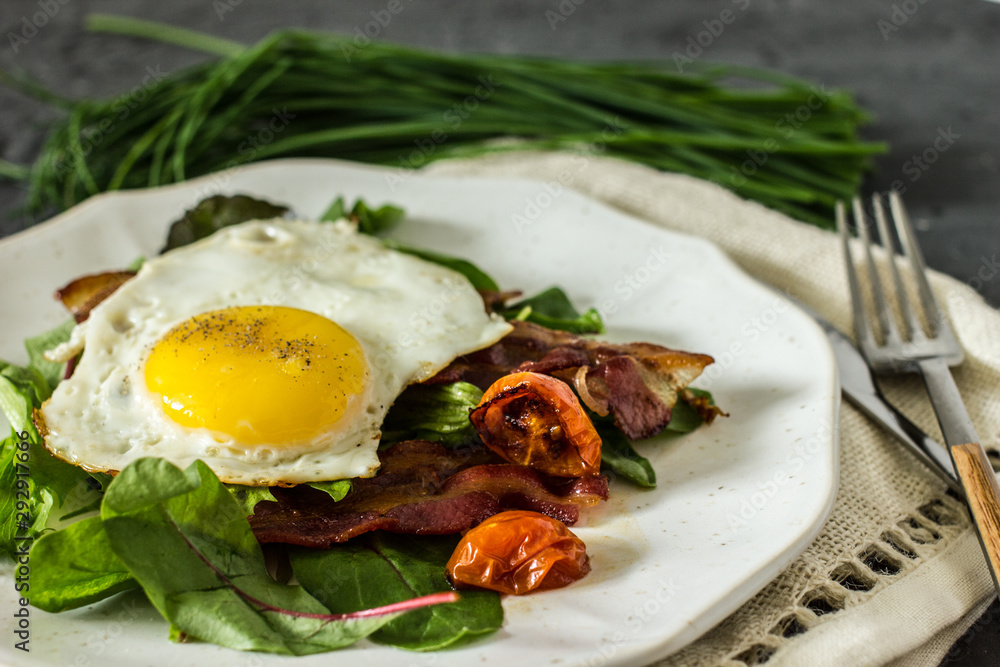 Appetizing breakfast with fried eggs and salad leaves. From above delicious meal with bacon egg tomatoes and green leaves served on white plate over gray textured background Appetizing breakfast with fried eggs and salad leaves. From above delicious meal with bacon egg tomatoes and green leaves served on white plate over gray textured background