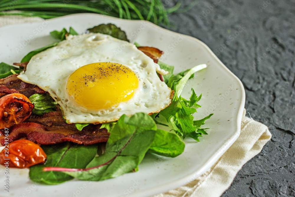 Appetizing breakfast with fried eggs and salad leaves. From above delicious meal with bacon egg tomatoes and green leaves served on white plate over gray textured background Appetizing breakfast with fried eggs and salad leaves. From above delicious meal with bacon egg tomatoes and green leaves served on white plate over gray textured background