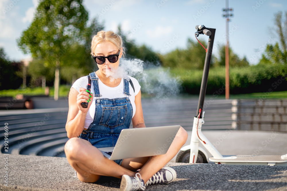 Modern woman smoking e-cigarette using her laptop computer while sitting at street with electric scooter