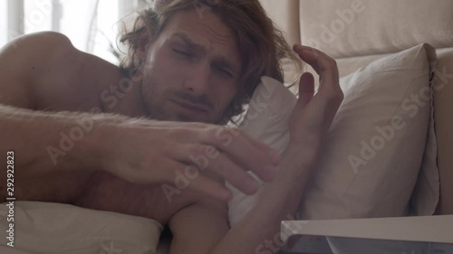 Slowmo shot of sleepy young man with stubble and long hair waking up in morning and checking time on his phone, then getting back to sleep photo