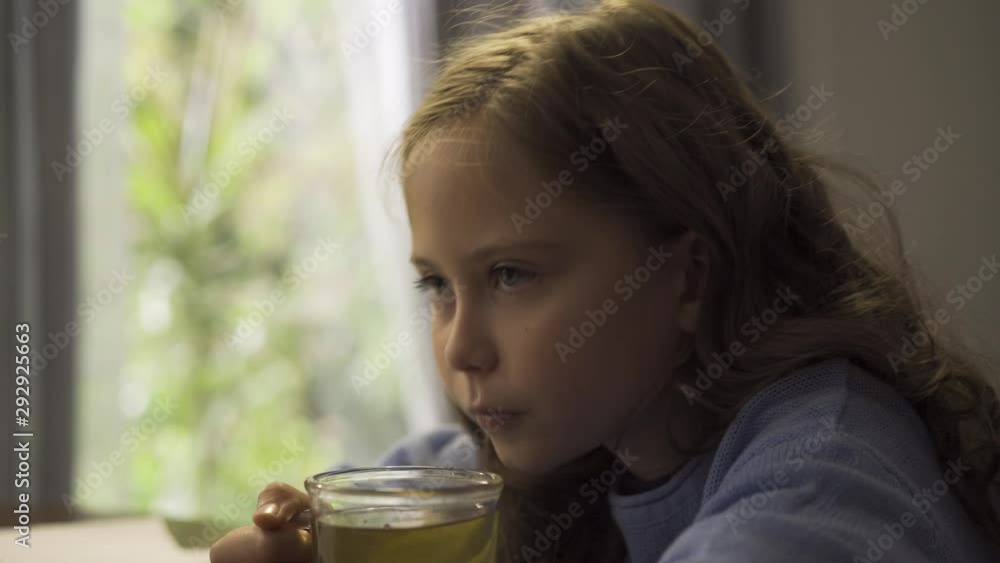 Pretty little girl drinking green tea sitting at the table in the kitchen. Caucasian child enjoying tea in the morning. Carefree childhood. Concept of healthcare and alternative medicine. Pretty little girl drinking green tea sitting at the table in the kitchen. Caucasian child enjoying tea in the morning. Carefree childhood. Concept of healthcare and alternative medicine.