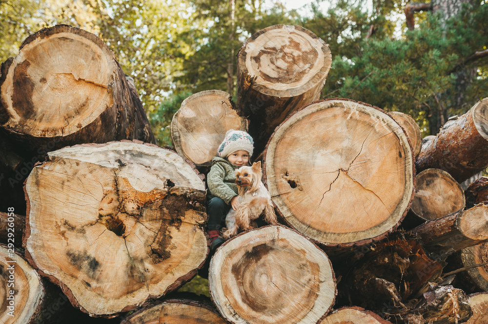 Happy little girl and small dog poses against the background of the big logs Happy little girl and small dog poses against the background of the big logs