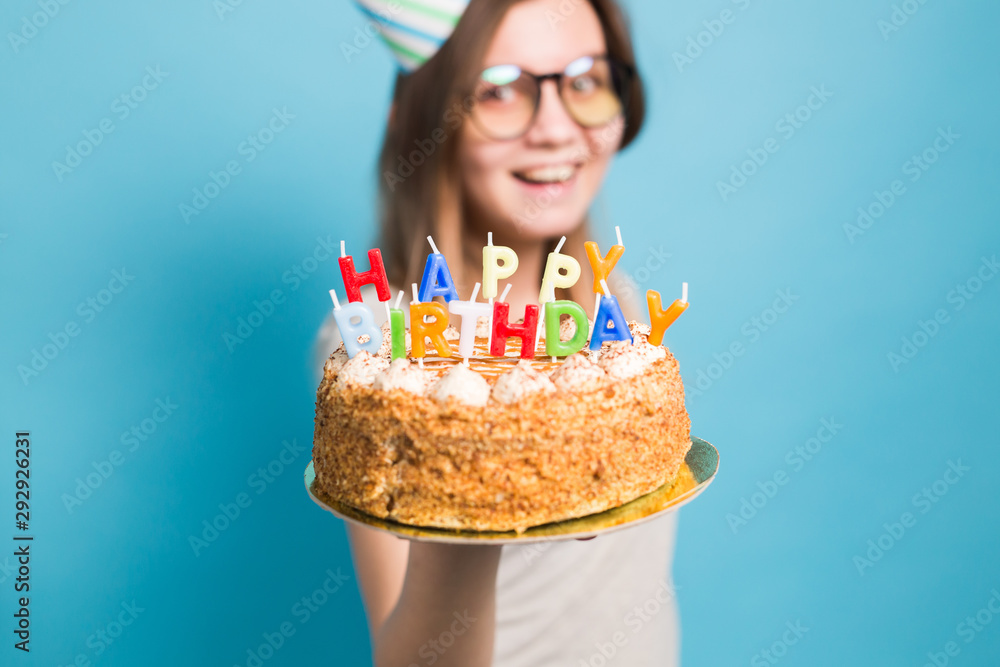 Charming merry crazy young girl student in congratulatory paper hat holding a happy birthday cake in her hands standing against a blue background. Advertising space. Charming merry crazy young girl student in congratulatory paper hat holding a happy birthday cake in her hands standing against a blue background. Advertising space.
