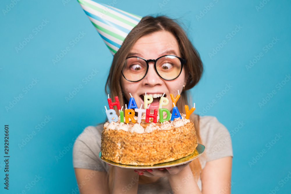 Φωτογραφία Close up funny positive girl in glasses and greeting paper hat holding a happy birthday cake in her hands standing on a blue background