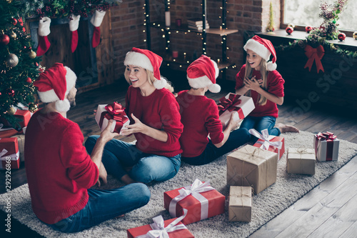 Photo of full family dad mom sister brother exchanging x-mas gifts sitting cozy floor carpet near decorated garland lights newyear tree indoors wear santa caps red jumpers