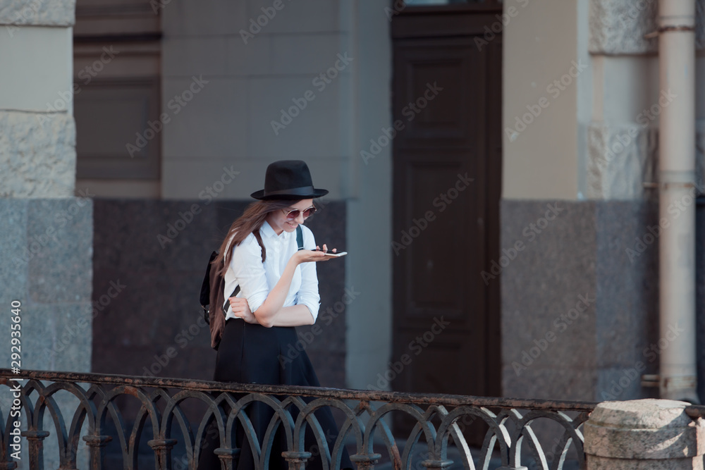 young woman in a hat and a white shirt walks in the city and uses a smartphone.