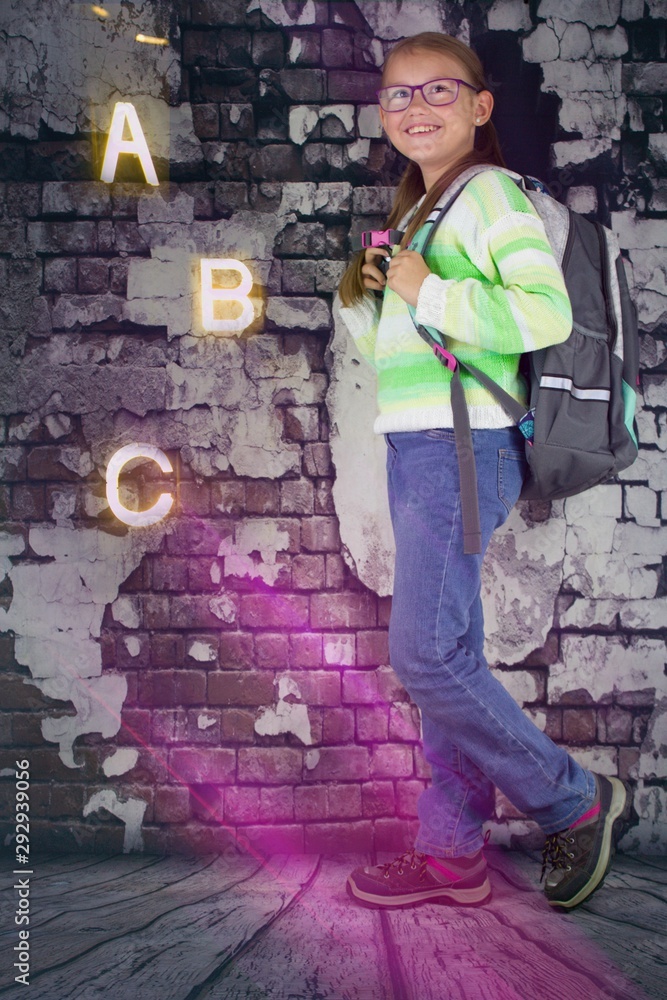 Obraz premium Girl with schoolbag on her back standing near old brick wall. Next to it are the letters A, B and C and the glare of light. Girl has glasses, striped sweater and blue jeans. Back to school concept