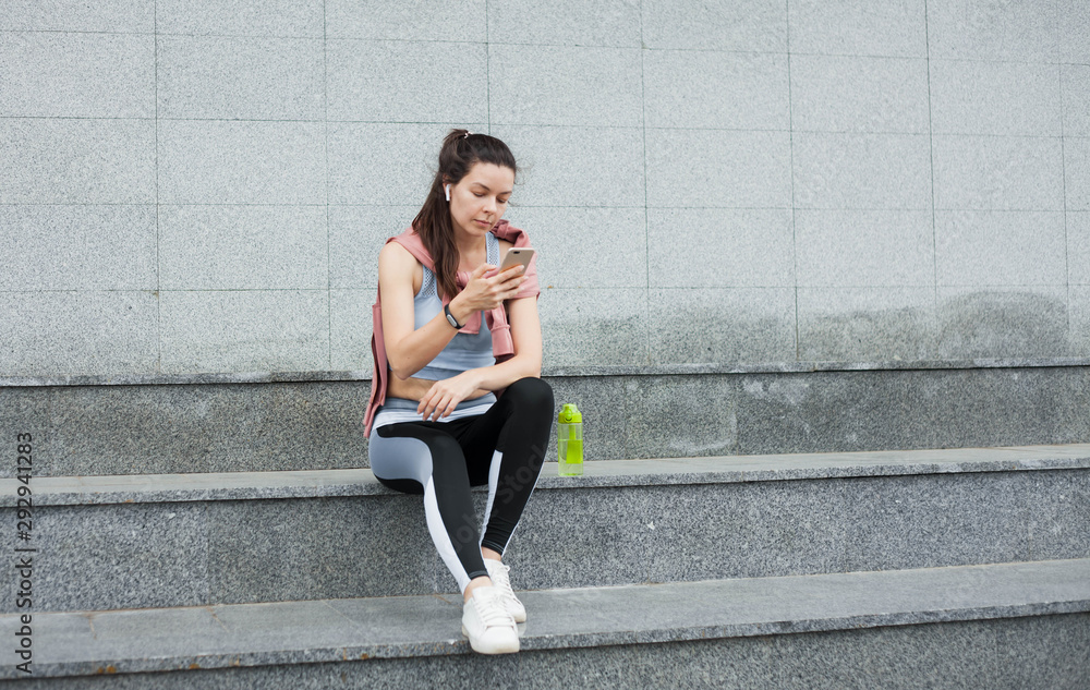 Fototapeta premium Athletic young woman in sports uniform sits, rests on the steps with smartphone and wireless headphones after a warm-up.