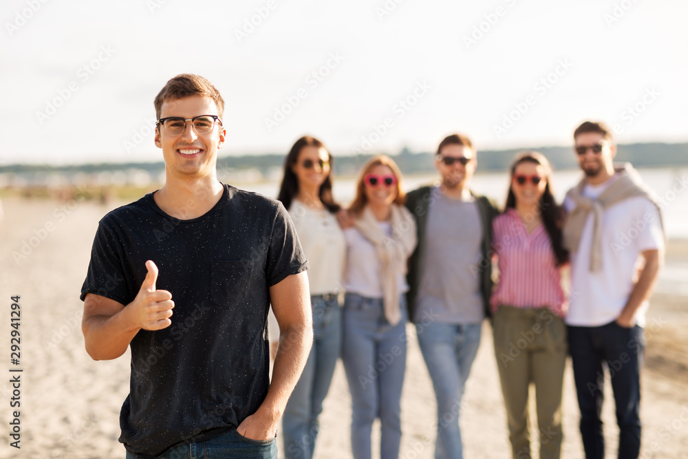 Obraz premium friendship, leisure and people concept - happy man with group of friends on beach in summer showing thumbs up