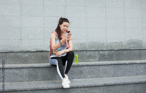 Athletic young woman in sports uniform sits, rests on the steps with smartphone and wireless headphones after a warm-up.