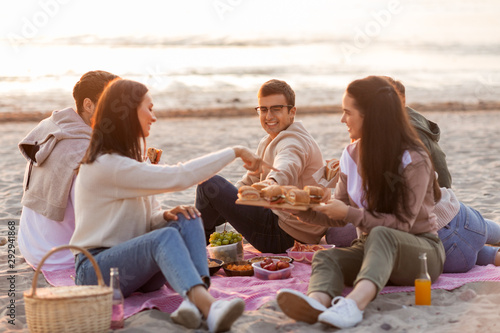 friendship  leisure and fast food concept - group of happy friends eating sandwiches or burgers at picnic on beach in summer