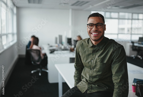 Portrait of a successful smiling young professional businessman sitting over desk in front of colleague working in background