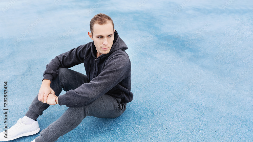 Young fit man in a gray sports suit sitting on the cover of the sports field resting after the morning run preparing for strength training. Healthy lifestyle and body concept. Copyspace