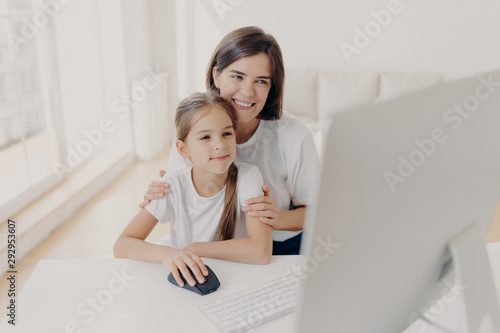 Happy affectionate mom embraces her small daughter, work together on modern computer, preapare presentation for lesson, look gladfully into monitor, spend leisure time at home, surf internet page. photo