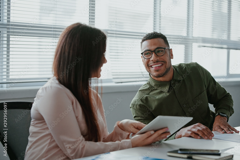 © StratfordProductions - Young businessman using a digital tablet to discuss information with her colleague in a modern office