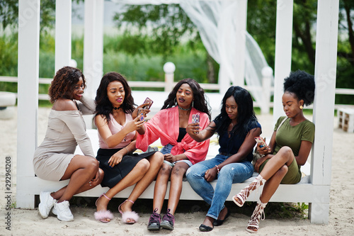 Group of five african american girls relaxing at beautiful poolside cabana beside luxury resort and looking at mobile phones.